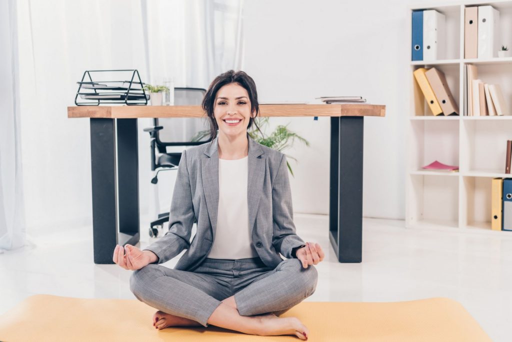 beautiful smiling businesswoman in suit sitting on fitness mat, looking at camera and meditating in